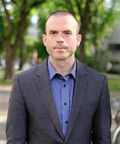 Man standing outside in a dark grey suit jacket and a collared blue shirt with trees in the background.