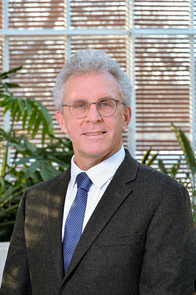 Man with white hair and light coloured glasses standing in an atrium in a black suit jacket, white shirt and blue tie. 