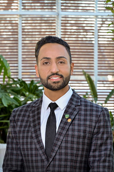 Man with dark hair and facial hair standing in an atrium wearing a dark colored tartan patterned suit jacket, white shirt and black tie. 