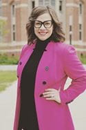 Woman with brunette hair, glasses, and a pink coat stands smiling in front of a brick buiding. 