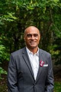 A man in a suit stands for a portrait, with trees in the background.