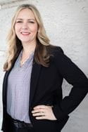 A woman with blonde hair, wearing a suit, poses with her hand on her hip in front of a white wall. 