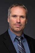 Man in blue suit and black jacket poses for a portrait. 