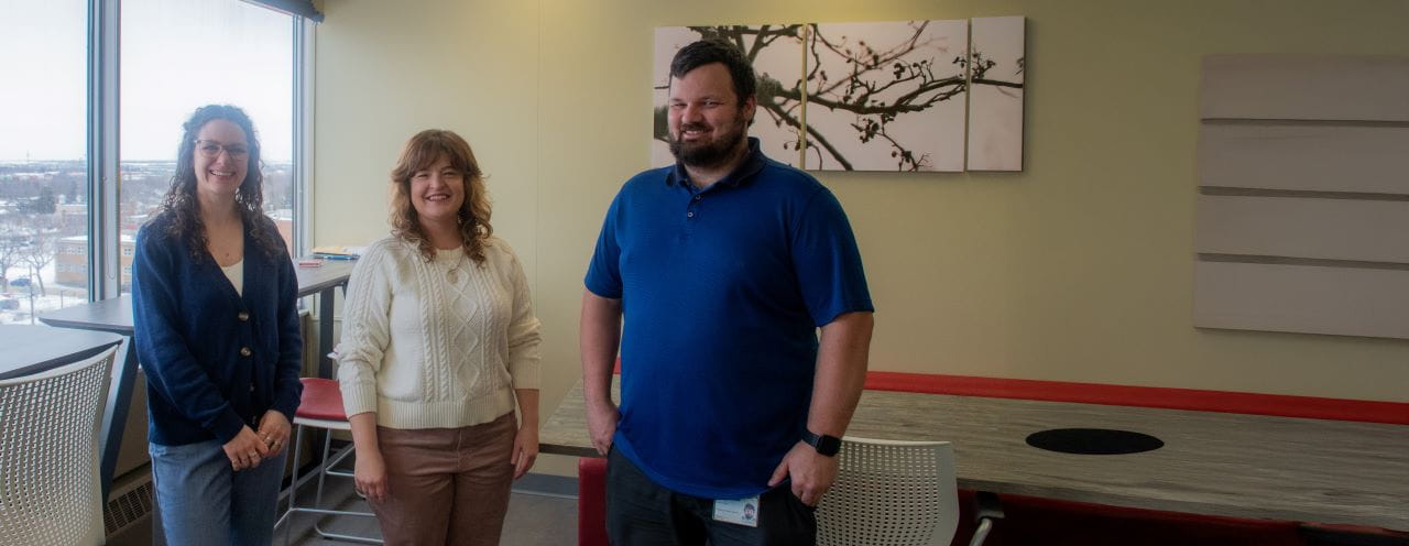 Three team members from Saskatchewan Accessibility Office standing in an office space, smiling at the camera. A large window and a modern table with chairs are in the background.