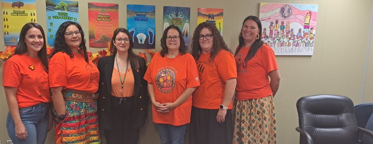 Six people celebrating Orange Shirt Day in front of a wall of Indigenous artwork.