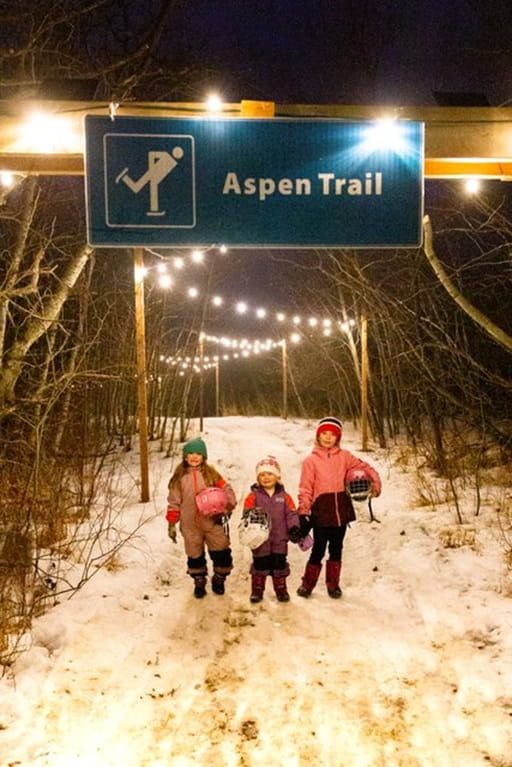 Three young girls stand in skates on an ice-skating loop underneath string lights at night.