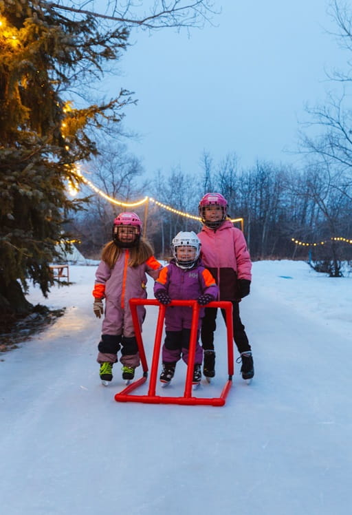 Three young girls on skates stand on an ice-skating loop underneath string lights at night.