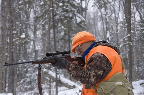 Senior man wearing a orange hat and orange vest holding a rifle in the woods. The ground is covered with white snow.  